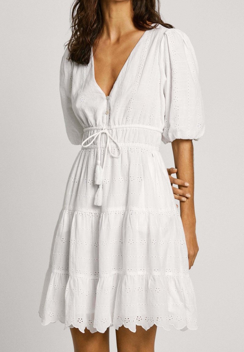 Femme portant une robe blanche en œillet avec manches bouffantes, col en V, détails de boutons, lien à pompons à la taille et ourlet festonné.
