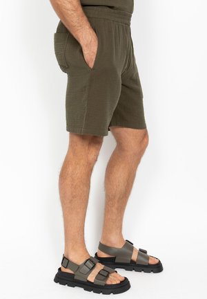 Homme portant un short texturé vert olive avec une ceinture élastique et des poches latérales, assorti à des sandales grises à boucle épaisse.