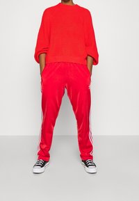 Rood gebreid vest met wijde mouwen, gecombineerd met glanzende rode trainingsbroek met witte zijstrepen. Zwarte sneakers maken de outfit compleet.