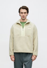 Κρεμ φλις πουλόβερ με μισό φερμουάρ μπροστά, που διαθέτει ανοιχτό πράσινο πάνελ και λογότυπο στο στήθος. Το ένδυμα έχει χαλαρή εφαρμογή και υφή επιφάνειας.