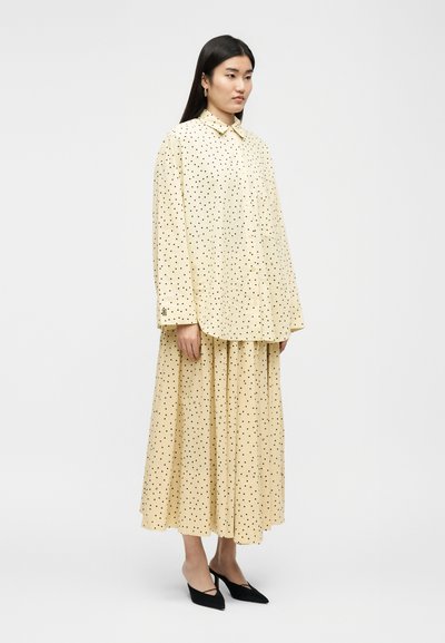 Γυναίκα που στέκεται, φορώντας μια χαλαρή μπεζ μπλούζα και μακρύ φούστα σε ταιριαστό χρώμα με μαύρες πουά, συνδυασμένη με μαύρες γόβες με μυτερή μύτη.