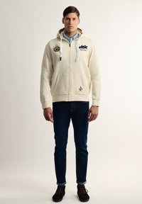 Sudadera crema con cremallera delantera, cordones, dos bolsillos y detalles de logo. Combinada con jeans oscuros y zapatos marrones.