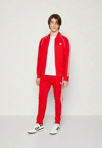 Jaunais cilvēks stāv, ģērbies sarkanā Adidas treniņtērpā ar baltām svītrām un baltās apavos, pret vienkāršu gaišu fonu.