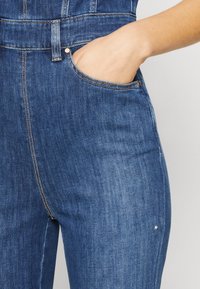 Pantalones de mezclilla con un lavado azul oscuro. Las características incluyen un bolsillo frontal, costuras en contraste y una cintura ajustada. Textura suave, diseño casual.