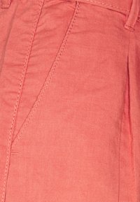 Yksityiskohta korallinvärisestä kankaasta, jossa on yksityiskohtaiset ompeleet ja vinosti oleva taskusauma vaatteen kohdalla.