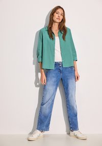 Camisa turquesa de botones y manga tres cuartos, combinada con jeans rectos de un azul claro y zapatillas blancas, sobre un fondo liso.