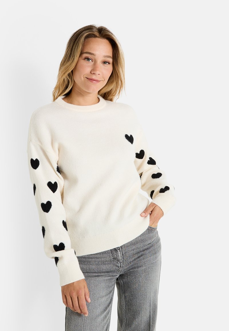 Suéter crema con motivos de corazones negros en las mangas. Cuello redondo, puños y dobladillo acanalados, textura suave. Combinado con jeans grises.