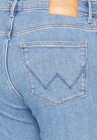 Jeans de mezclilla azul claro con un bolsillo trasero que presenta un patrón de costura en zigzag y una etiqueta de cuero marrón con la inscripción "Wrangler" en relieve.