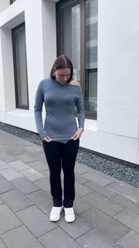 Pilkas ilgomis rankovėmis viršus su off-shoulder dizainu, derinamas su juodomis kelnėmis ir baltomis sportinėmis bateliais, pozuojantis akmeniniuose takeliuose.