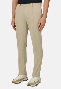 Pantalon beige ajusté avec coutures avant porté avec des baskets épaisses beige et blanches sur fond blanc.