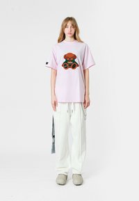Una camiseta rosa de gran tamaño presenta un gráfico de un oso de peluche en marrón y verde. Combinada con pantalones blancos y zapatos grises texturizados.