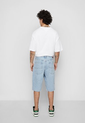 Chemise blanche à manches courtes associée à un short en denim bleu clair. L'individu porte des baskets montantes noires et vertes avec des semelles blanches.