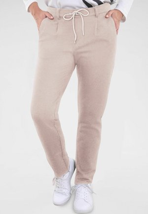 Beige, schmal zulaufende Hose aus weichem Stoff mit einem Kordelzugbund. Mit Fronttaschen und glatter Textur. Kombiniert mit weißen Sneakern.