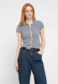 Kortærmet bluse med krave i sort og hvid gingham-mønster, kombineret med højtaljede mørke denimjeans, fastgjort med et sort bælte.