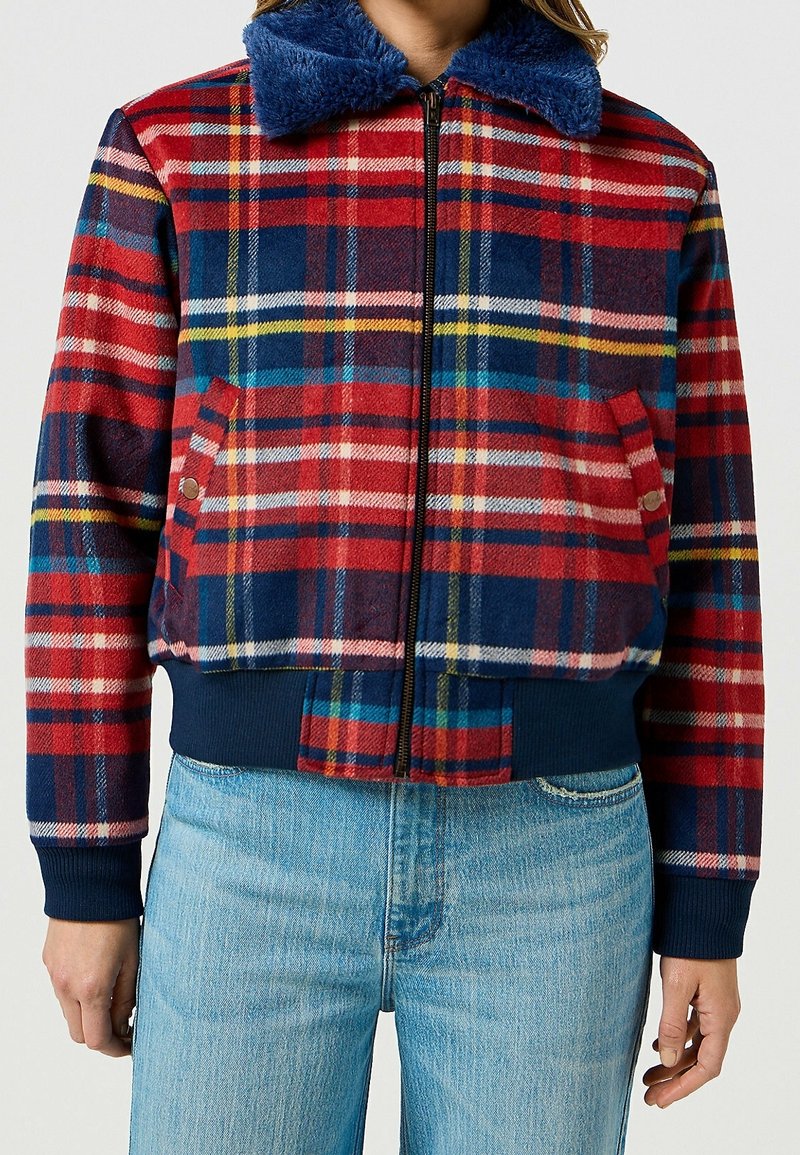 Veste bomber à carreaux rouge, bleu et jaune avec un col en fausse fourrure. Elle comprend des poignets et un ourlet côtelés, deux poches latérales et une fermeture éclair.