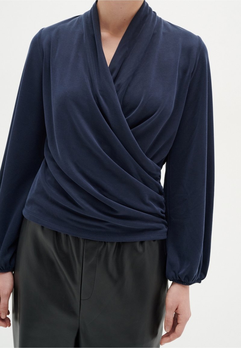 Femme portant un top drapé à manches longues bleu marine associé à un pantalon en cuir noir taille haute, debout devant un fond uni.