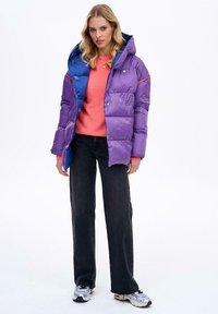 Fialová nafukovacia bunda s modrou kapucňou, s prídavkom zipsu, patentiek a dvoma vreckami. Nosená cez ružový sveter a čierne džínsy.