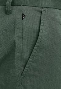 Gröna tygbyxor har ett texturerat mönster, sidfickor och en liten svart triangulär logotyp fram.