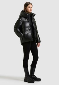Giacca nera imbottita con un grande cappuccio, texture liscia e due tasche frontali. Indossata con leggings neri e stivali da combattimento con lacci.
