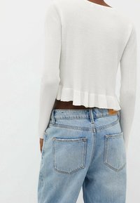 Person, der helle blaue High-Waist-Jeans und ein weißes, geripptes langärmliges Oberteil mit rüschengesäumtem Saum trägt, von hinten betrachtet.