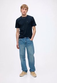 Joven de pie con camiseta negra, vaqueros azul claro, zapatillas beige y un collar de cadena plateada frente a un fondo blanco liso.