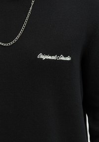 Black sweater made of soft fabric features a classic crew neckline and embroidered "Originals Studio" logo in white. Chain detail visible.