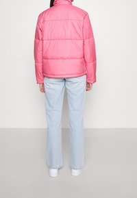 Puffjacka i glansig rosa, med hög krage, horisontella sömmar och en avslappnad passform, kombinerad med ljusblå utsvängda jeans och vita sneakers.