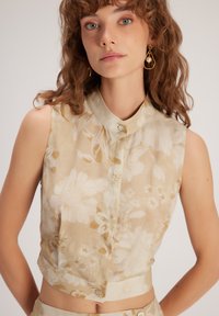 Top corto sin mangas floral en beige con patrones crema. Presenta un cuello con solapa y un cierre de botones en la parte delantera.