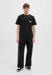 Schwarzes Baumwoll-T-Shirt mit kurzen Ärmeln und weißem Logo auf der linken Brust, kombiniert mit lockeren dunkelgrauen Jeans und schwarzen Sneakers.