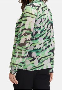 Donna che indossa una blusa a maniche lunghe, semitrasparente, con motivi astratti di foglie verdi, nere e bianche e polsini arricciati, abbinata a pantaloni neri.