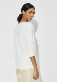 Biała, luźna bluzka z rękawami do 3/4, okrągłym dekoltem, miękką fakturą i gładką sylwetką. Noszona z jasnymi spodniami.