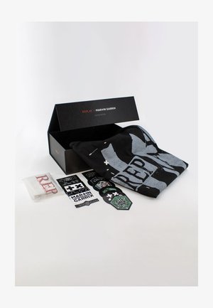 Sort t-shirt med stort gråt grafisk design, pakket i en sort æske. Inkluderer klistermærker og et hvidt stof med rød tekst. Begrænset udgave varer.