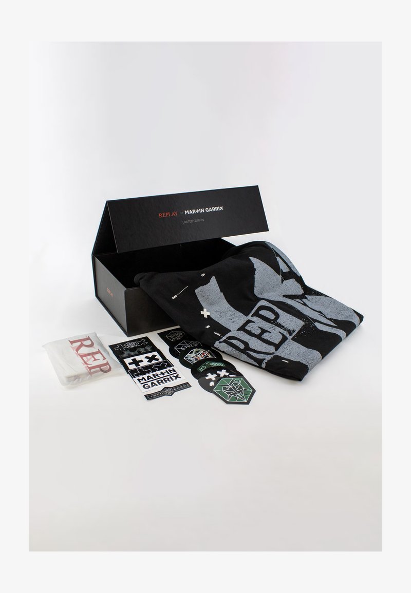 T-shirt noir avec un grand graphisme gris, emballé dans une boîte noire. Comprend des autocollants et un chiffon blanc avec un texte rouge. Articles en édition limitée.