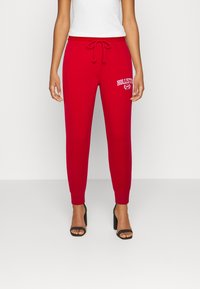 Röda joggarbyxor i mjukt tyg, med dragsko i midjan och en vit "Hollister"-logotyp på fram vänster. Åtsittande muddar vid anklarna.