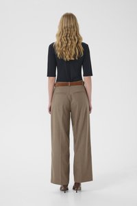 Naine laineliste blondide juustega, kandes musta küünarnukivarrukatega toppi, pruune laia säärega pükse, pruuni vööd ja pruune kontsakingi, seistes seljaga vaataja poole.