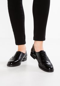 Des chaussures brogue en cuir verni noir portées avec un pantalon noir ajusté sur un fond de sol blanc.
