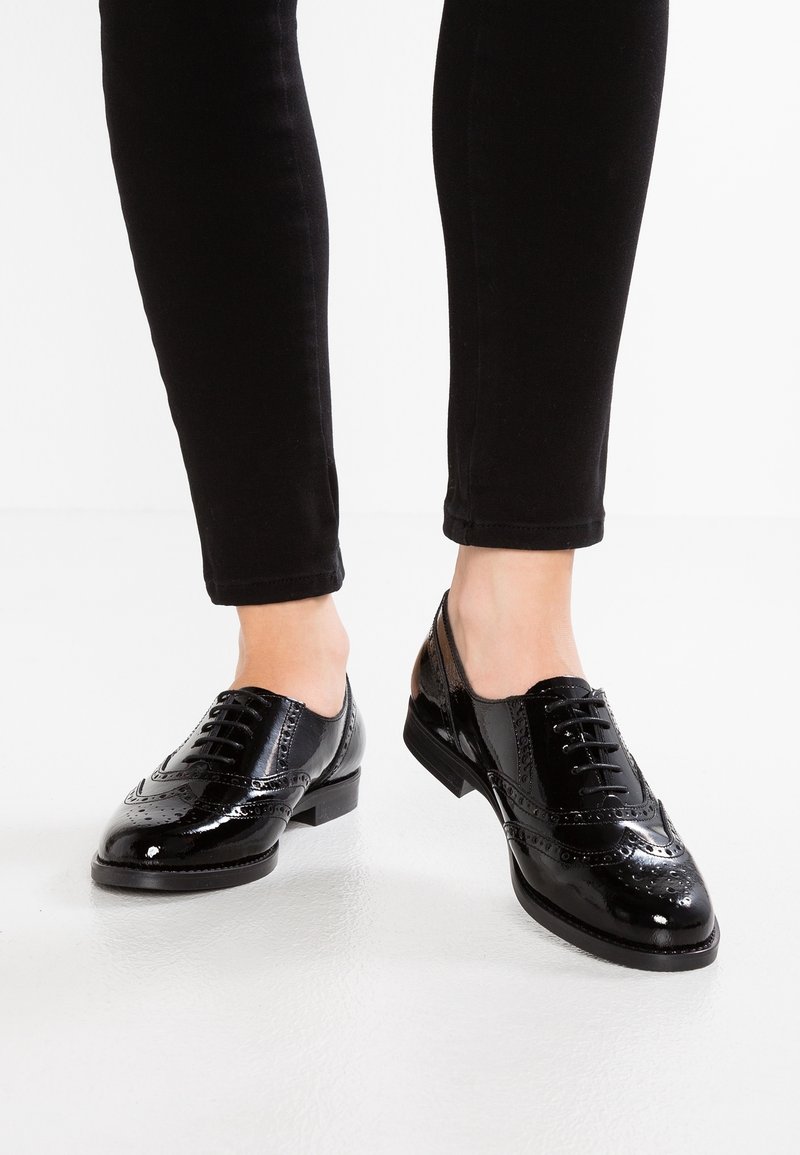 Des chaussures brogue en cuir verni noir portées avec un pantalon noir ajusté sur un fond de sol blanc.