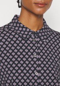 Gros plan sur une personne portant une chemise noire à col avec un motif géométrique blanc et violet, boutonnée jusqu'au cou.