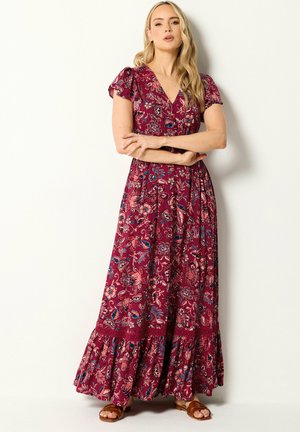 Vrouw in een bordeaux rode maxidress met korte pofmouwen, V-hals en ruchtrand, staand met gekruiste armen, draagt bruine sandalen.