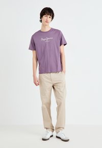 Purple cotton T-shirt with rounded neck, featuring white "Pepe Jeans London" text, paired with beige trousers and white sneakers.