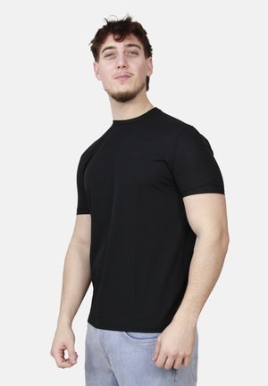 Giovane uomo con capelli corti e ricci e leggera barba, indossa una maglietta nera attillata a maniche corte e jeans azzurri, in piedi davanti a uno sfondo neutro.