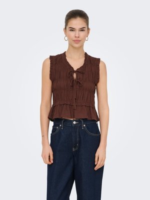 Ung kvinne med trukket hår bakover, iført gull ørepynt i form av hoops, en brun ermeløs topp med rynker og knyter på sidene, samt høytlivs mørkeblå jeans.