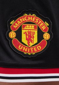 Blason du club de football Manchester United brodé sur tissu noir, représentant un diable rouge et deux ballons de football, au-dessus de bandes rouges, blanches et noires.