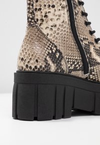 Stivaletto alla caviglia con motivo pitonato, superiore beige testurizzato, lacci neri e suola nera spessa e rigata. Zip laterale per una facile vestibilità.