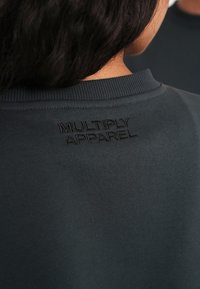 Sweatshirt gris foncé avec un col côtelé et le texte en relief "MULTIPLY APPAREL" au dos. Tissu en coton doux avec une texture lisse.