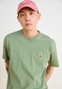 Green cotton t-shirt with a patch pocket featuring a logo. Worn with a light pink cap. The fabric appears soft with a relaxed fit.