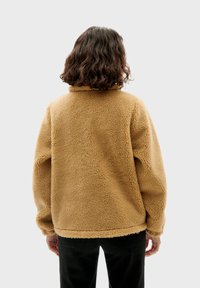 Beige fleece jacket met hoge kraag, zachte textuur en verlaagde schouders, gecombineerd met zwarte broek, gezien vanaf de achterkant.