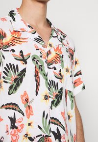 Chemise à manches courtes présentant un fond blanc avec des motifs floraux et d'oiseaux vibrants en orange, vert, noir et jaune, avec un col et des boutons.
