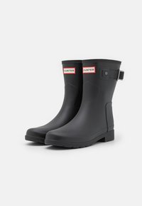 Botas de lluvia de goma negra con un acabado liso, ligero tacón, detalle de hebilla en el lateral y logo blanco "HUNTER" en la parte frontal.