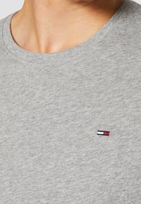 Primo piano di una persona che indossa una maglietta a girocollo grigia chiara con un piccolo logo rettangolare ricamato rosso, bianco e blu navy sul petto.